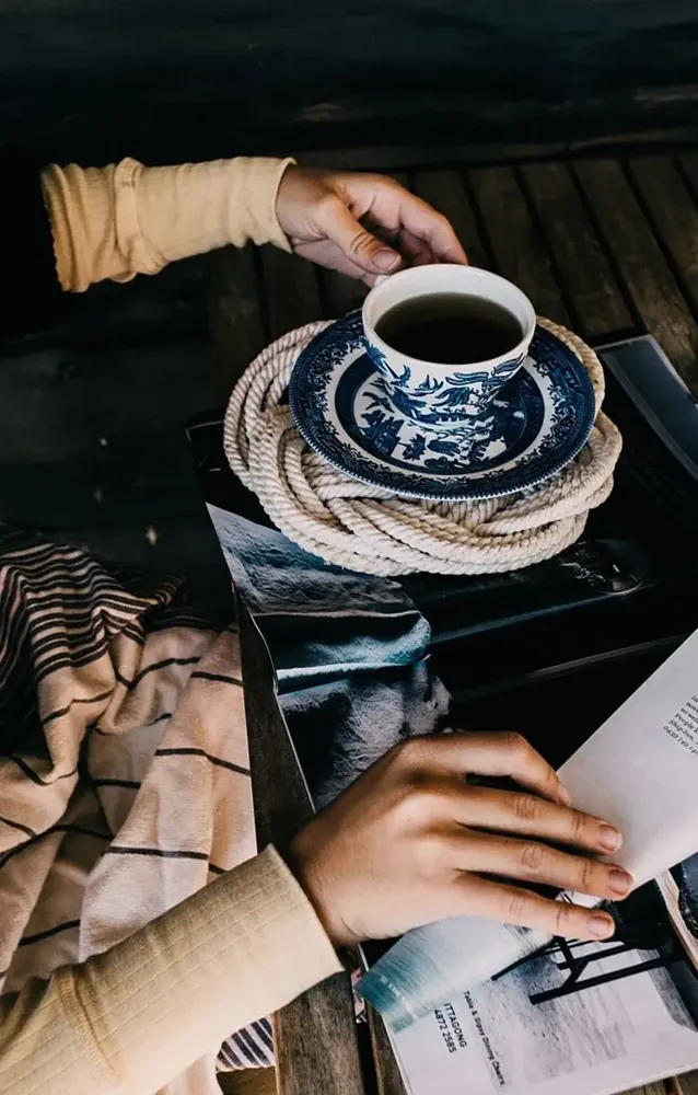 Hands holding a patterned cup of coffee beside open papers on a dark table