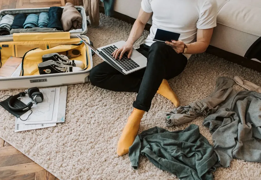 Man sitting on a rug packing a suitcase while using a laptop
