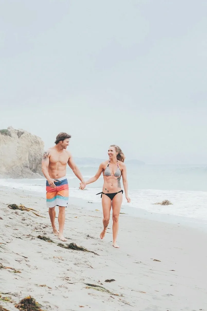 Couple in swimwear walking hand in hand along a pale beach