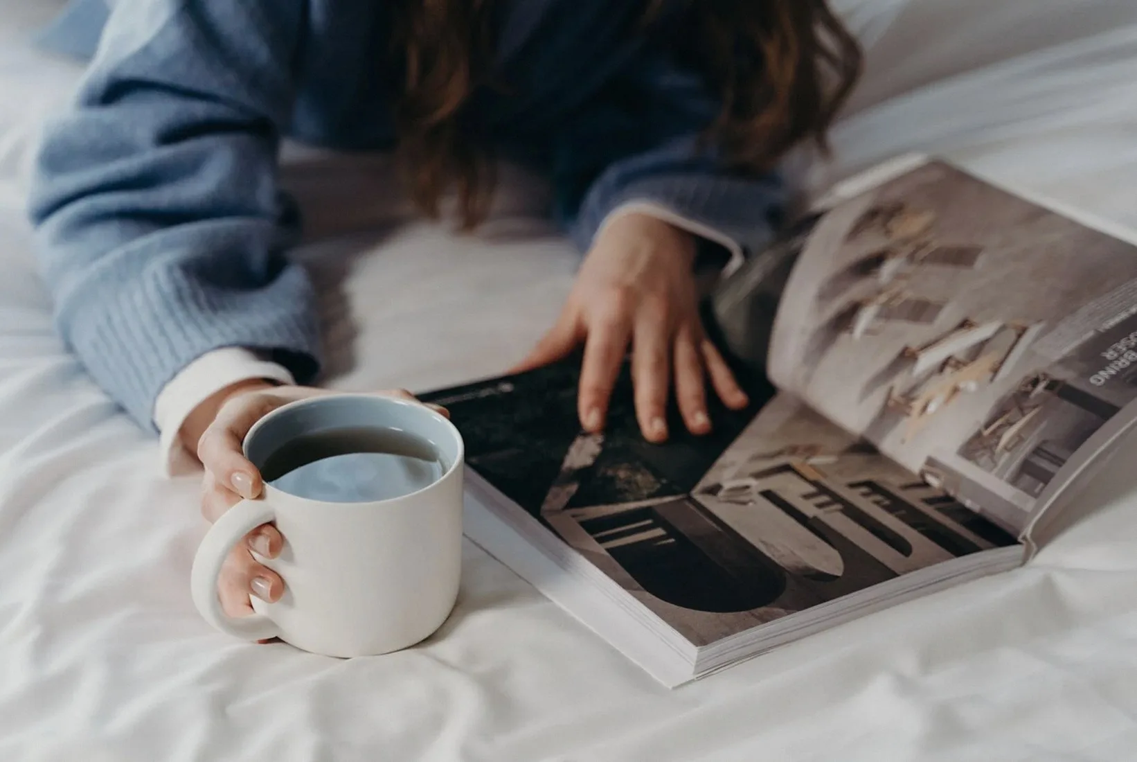 Person reading an open magazine in bed with a mug of coffee